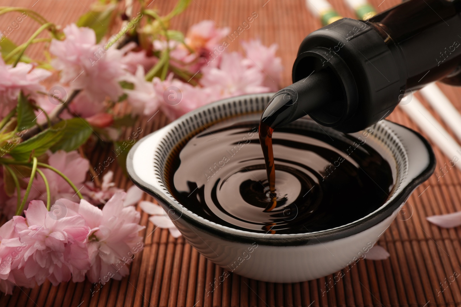 Photo of Pouring soy sauce into bowl and beautiful blossoming branch on bamboo mat, closeup