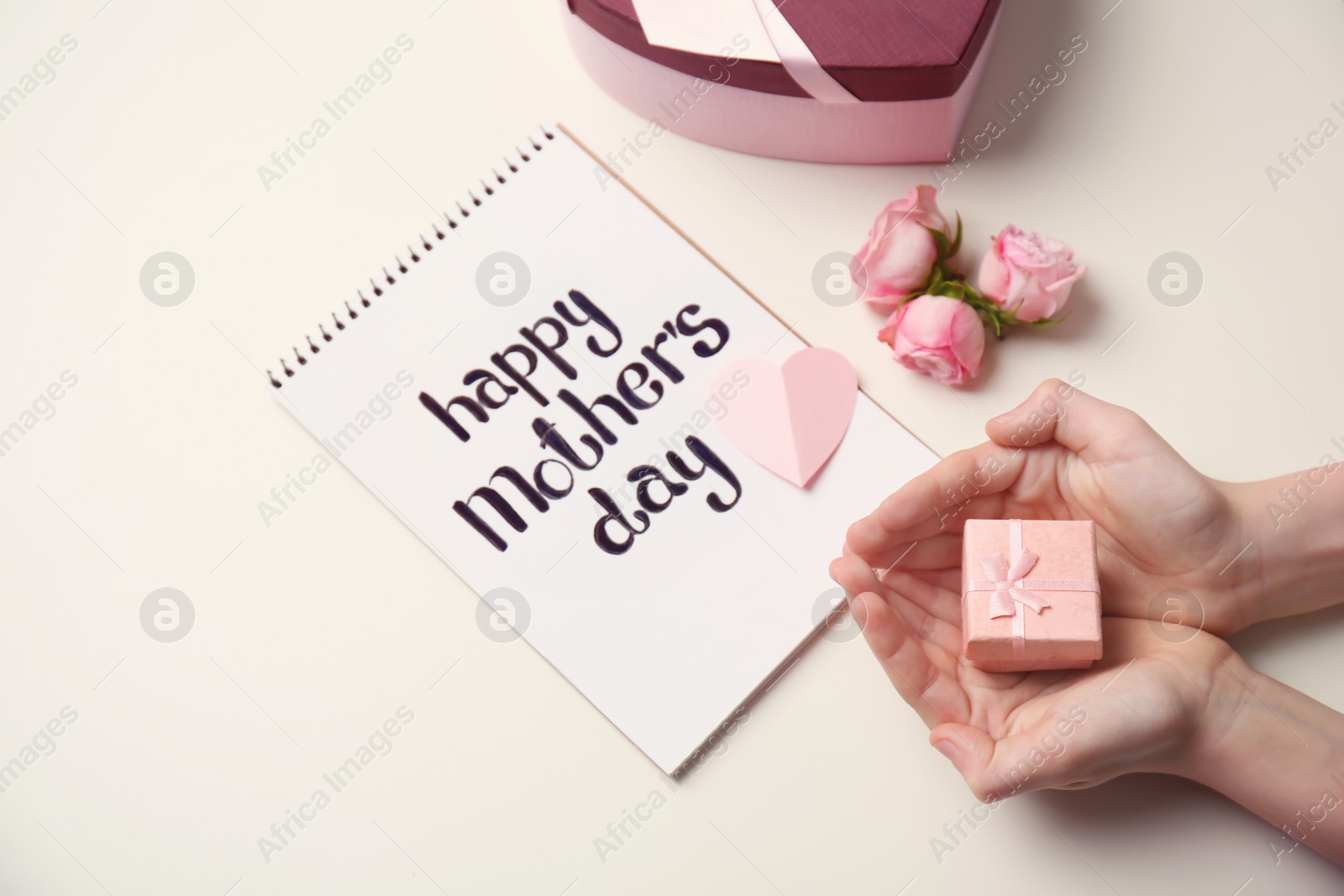 Photo of Little child holding gift box near notebook with words HAPPY MOTHER'S DAY on light background