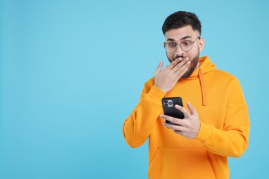 Shocked young man using smartphone on light blue background, space for text