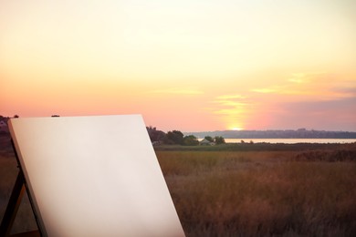 Image of Wooden easel with blank canvas in field at sunrise. Space for text