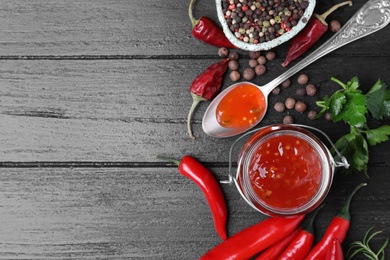 Jar and spoon of hot chili sauce with ingredients on wooden background, flat lay. Space for text