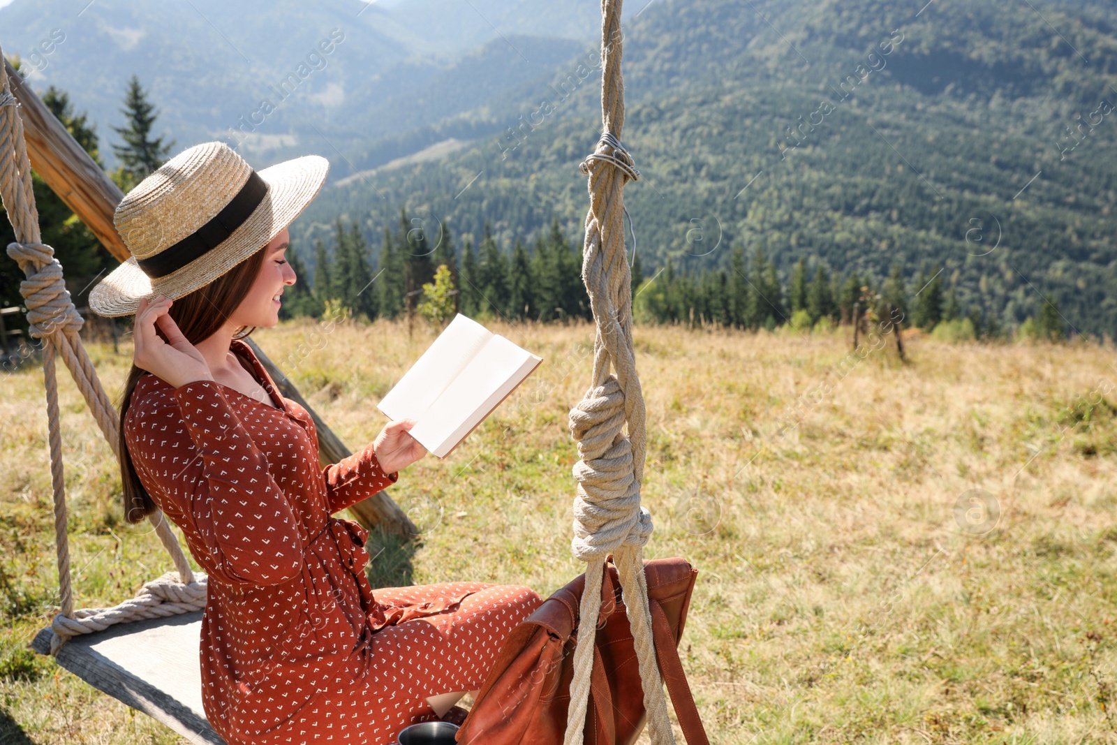 Photo of Beautiful young woman reading book on wooden swing in mountains, space for text