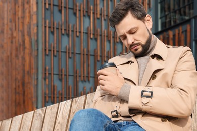 Tired man with cup of coffee sleeping while sitting on wooden bench outdoors. Space for text