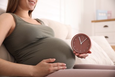Photo of Young pregnant woman holding clock near her belly at home, closeup. Time to give birth