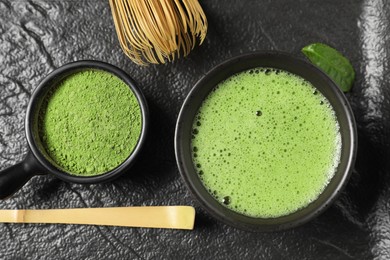 Photo of Cup of fresh matcha tea, bamboo whisk, spoon and green powder on black tray, flat lay