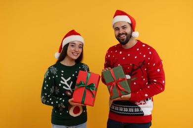 Photo of Happy young couple in Christmas sweaters and Santa hats with gifts on orange background