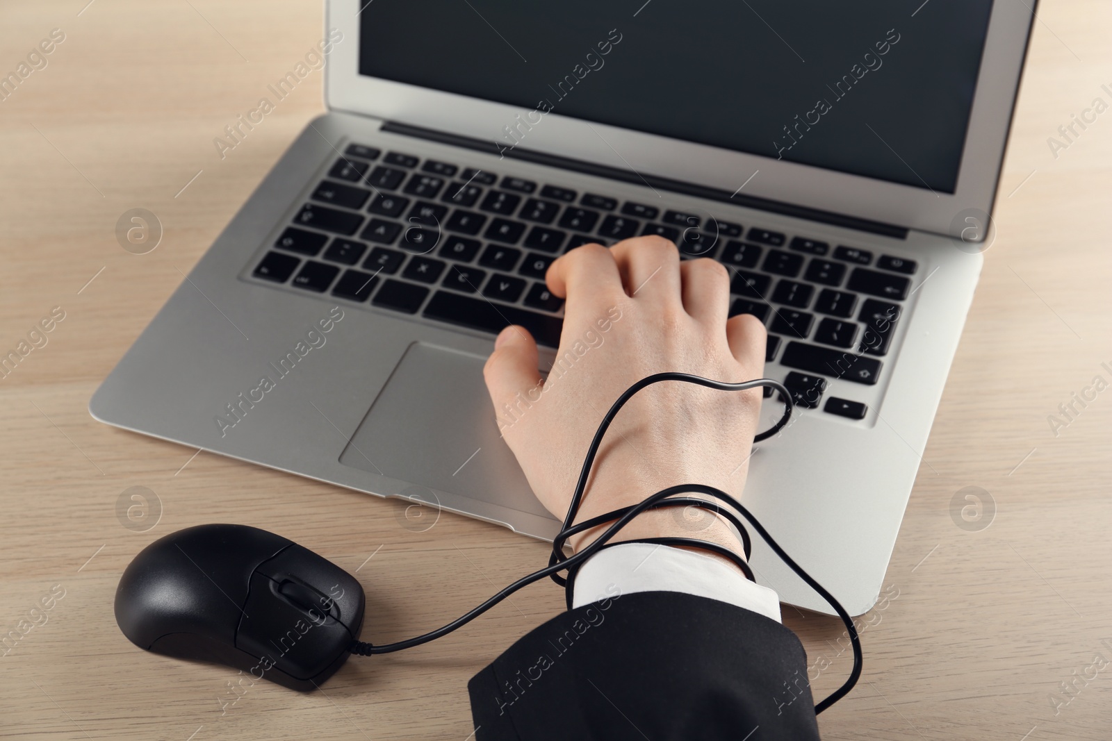 Photo of Internet addiction. Closeup view of man typing on laptop at wooden table, hand tied with computer mouse cable