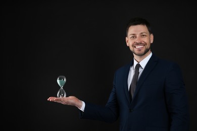 Happy businessman holding hourglass on black background. Time management