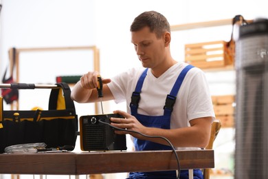 Photo of Professional technician repairing electric fan heater with screwdriver at table indoors