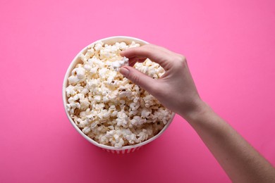 Woman taking delicious popcorn from paper bucket on pink background, top view