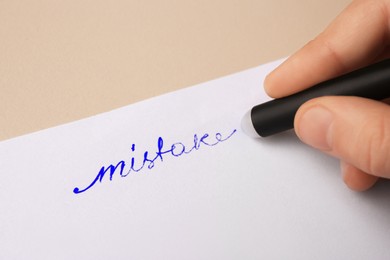 Child erasing word Mistake written with erasable pen on paper sheet against beige background, closeup