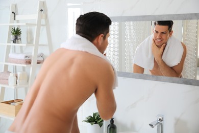 Photo of Handsome man touching his smooth face after shaving near mirror in bathroom