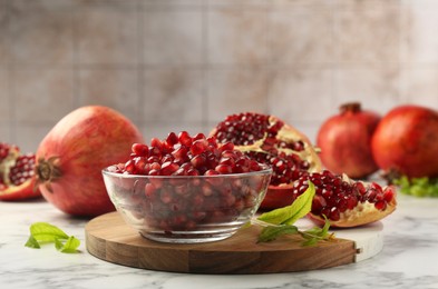 Photo of Ripe juicy pomegranate grains in bowl and green leaves on white marble table