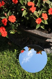 Photo of Round mirror on green grass reflecting sky and beautiful flowers, above view
