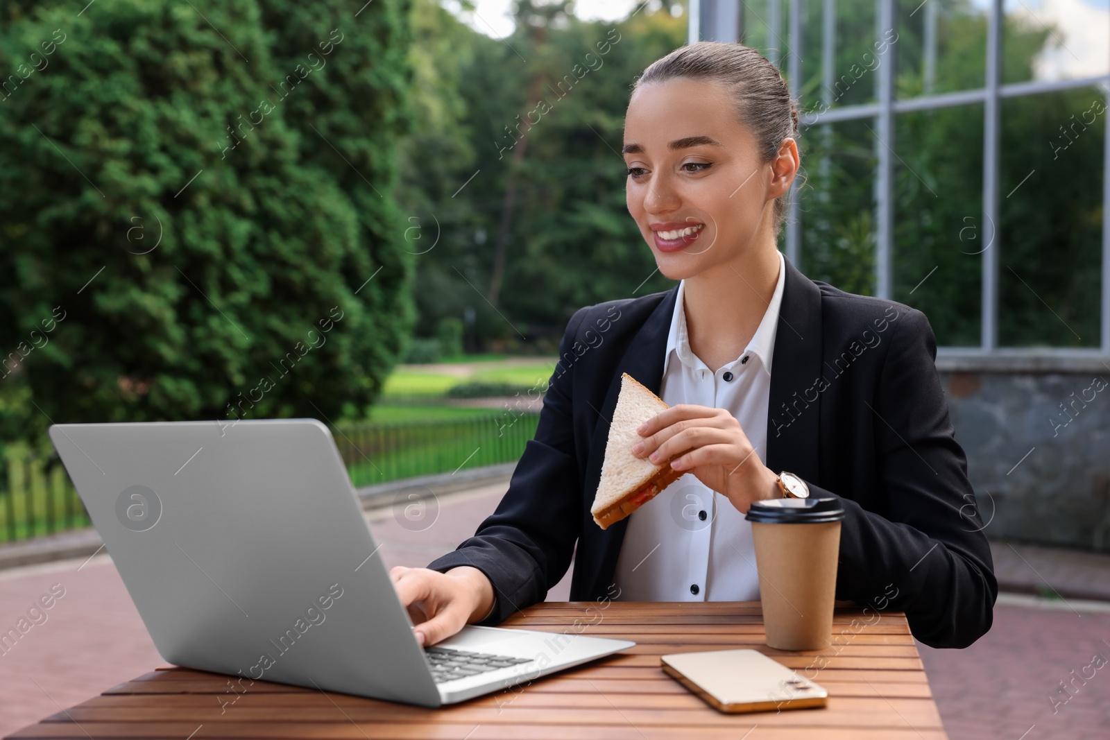 Photo of Happy businesswoman with sandwich using laptop while having lunch at wooden table outdoors
