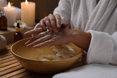 Photo of Woman applying scrub onto her hands in spa, closeup. Bowl of water and flowers on wooden table