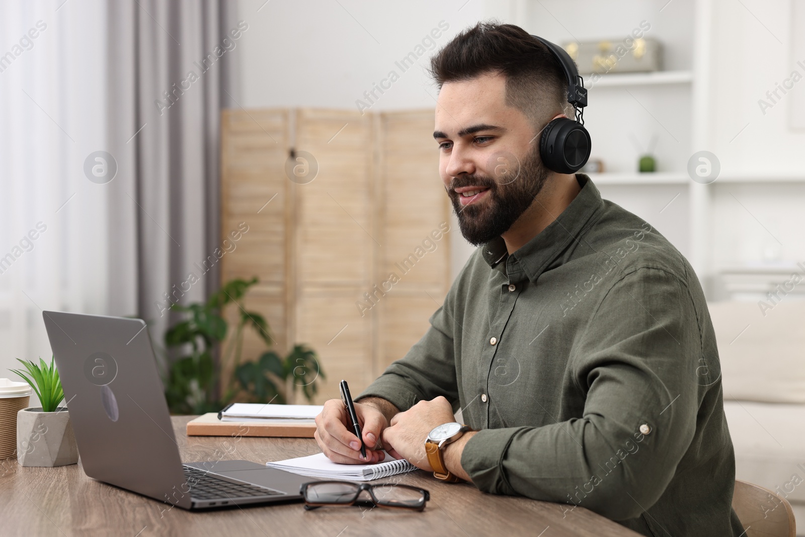 Photo of E-learning. Young man taking notes during online lesson at wooden table indoors