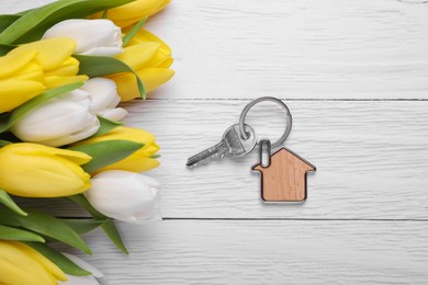 Beautiful spring flowers and key with trinket in shape of house on white wooden table, flat lay