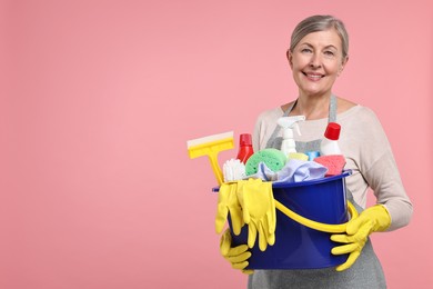 Photo of Happy housewife holding bucket with cleaning supplies on pink background, space for text