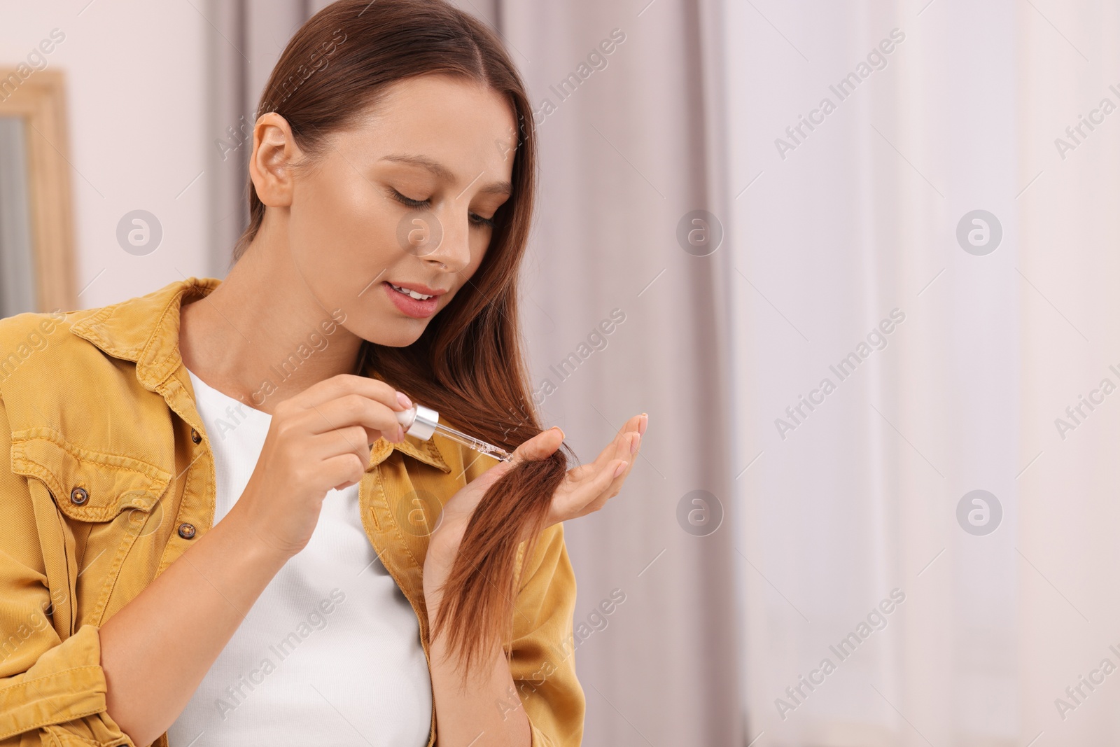 Photo of Beautiful woman applying serum onto hair indoors, space for text