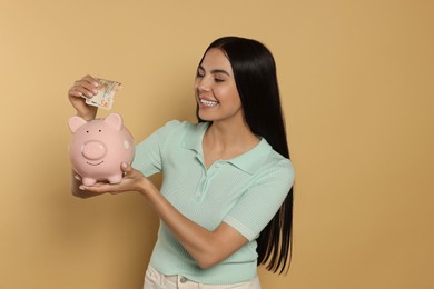 Photo of Happy young woman putting money into piggy bank on beige background