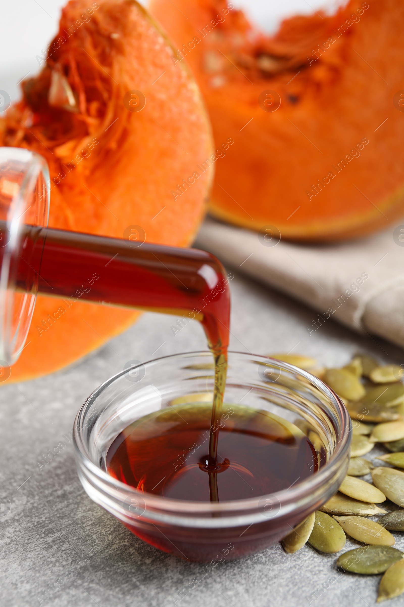 Photo of Pouring pumpkin oil into bowl and seeds on light grey table, closeup