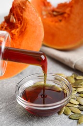 Photo of Pouring pumpkin oil into bowl and seeds on light grey table, closeup