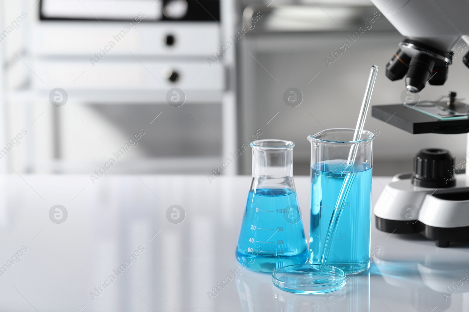 Photo of Different laboratory glassware with light blue liquid near microscope on table, space for text