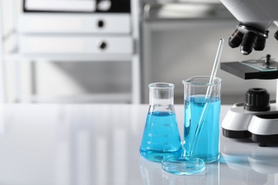 Photo of Different laboratory glassware with light blue liquid near microscope on table, space for text