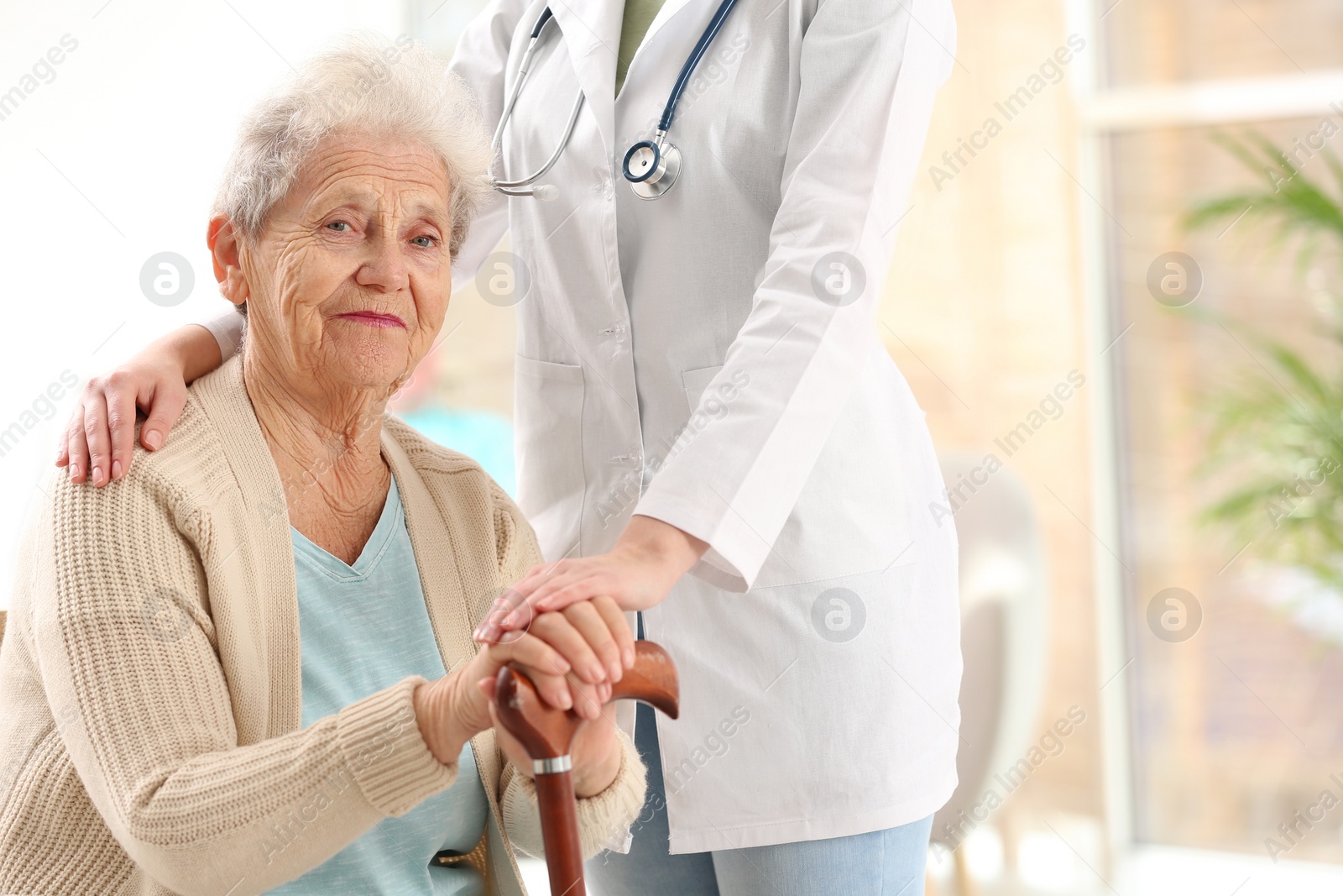 Photo of Nurse assisting elderly woman with cane indoors. Space for text