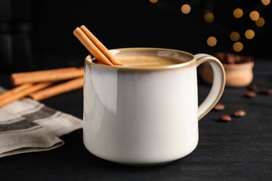 Cup of hot coffee with aromatic cinnamon on black wooden table against blurred lights