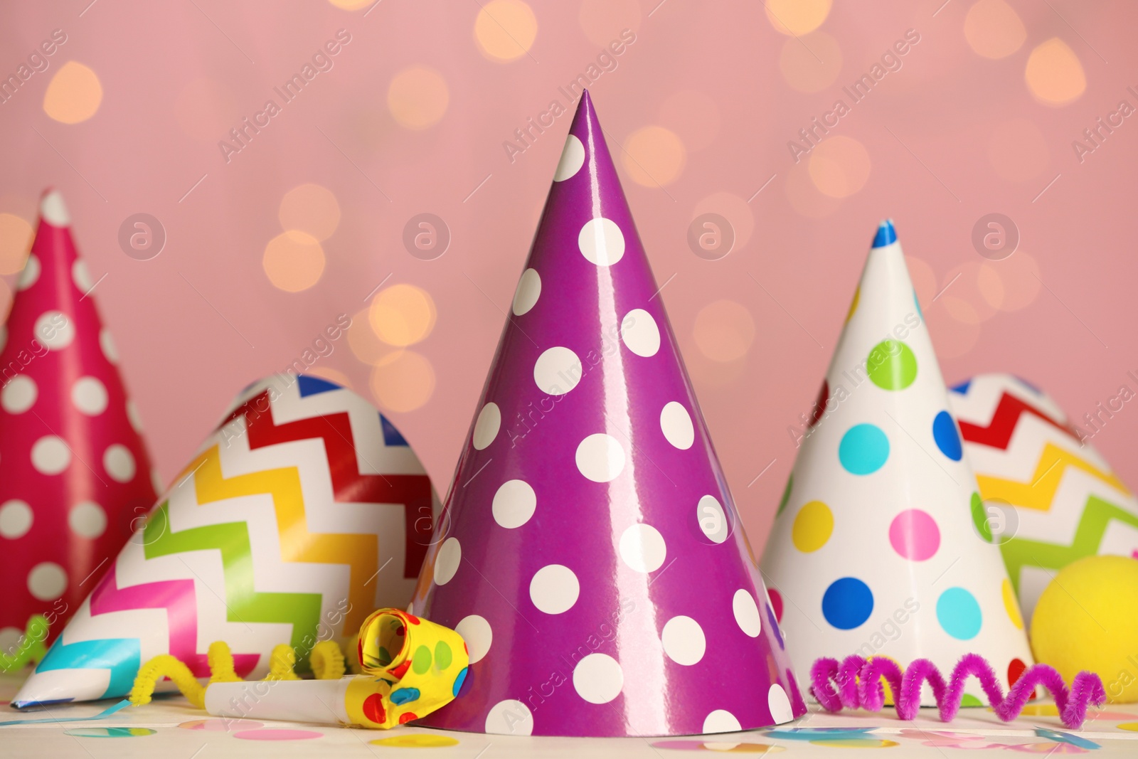 Photo of Colorful party hats and festive items on white table against pink background with blurred lights