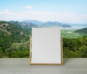 Image of Wooden easel with blank canvas on table and beautiful mountain landscape. Space for text 