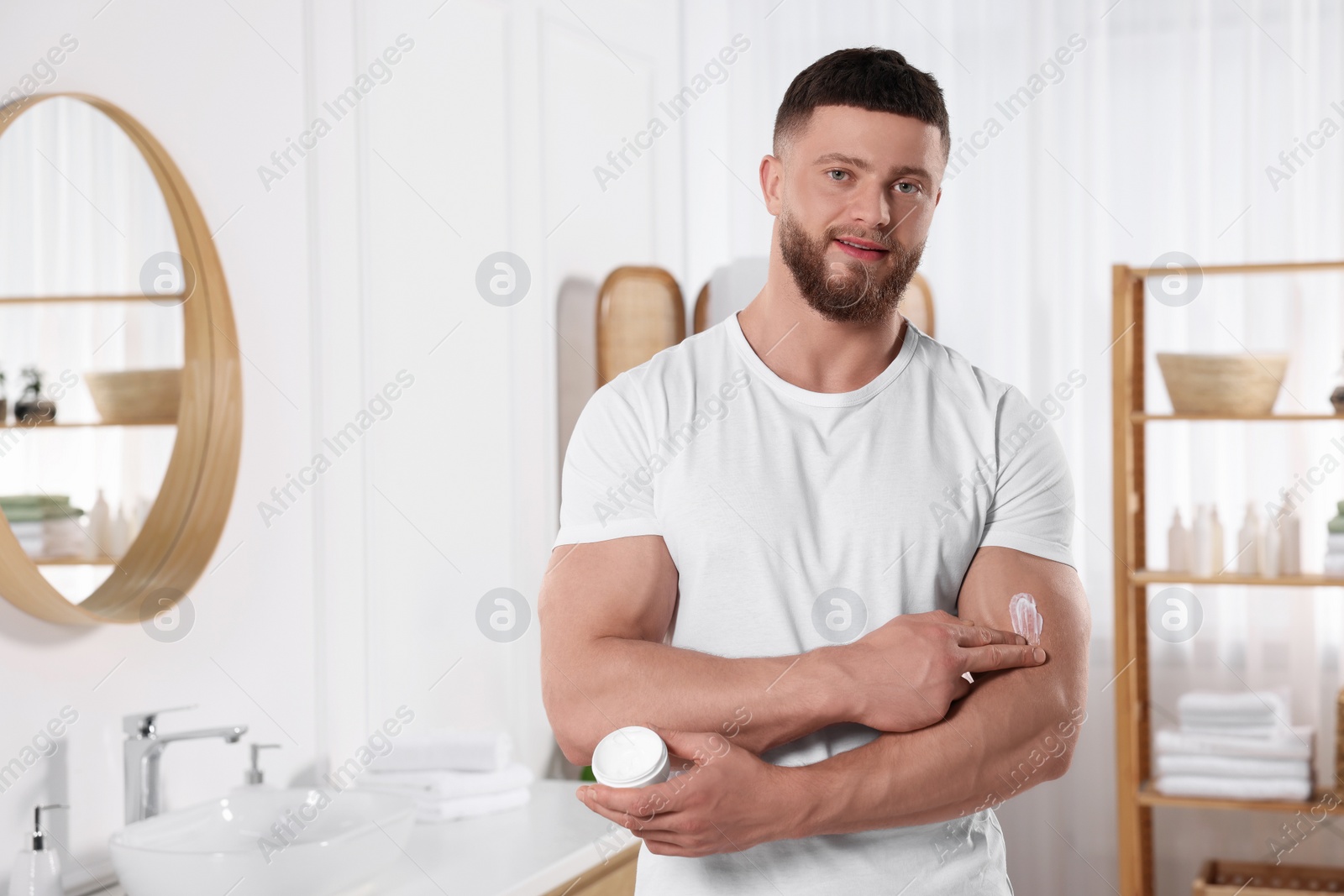 Photo of Handsome man applying body cream onto his arm in bathroom. Space for text