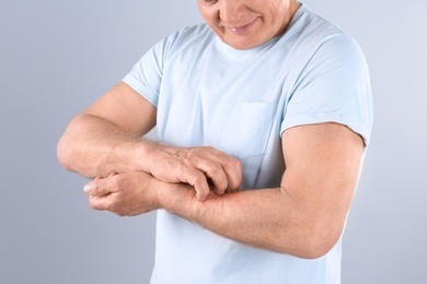 Senior man scratching forearm on grey background, closeup. Allergy symptom