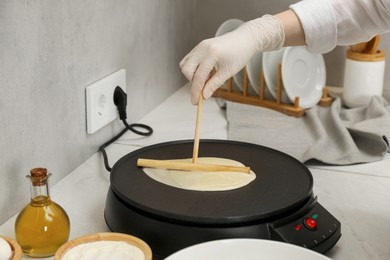 Photo of Woman cooking delicious crepe on electric pancake maker at white marble table in kitchen, closeup