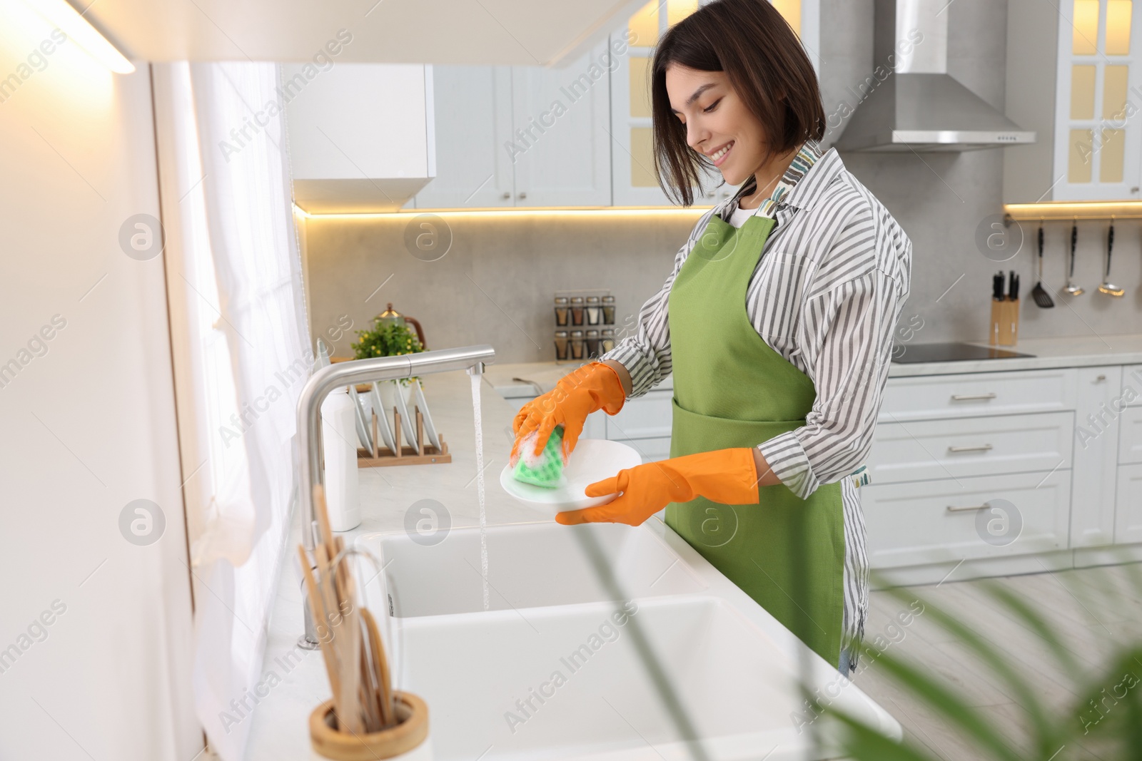 Photo of Happy young woman washing plate above sink in modern kitchen