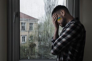 Upset man with picture of Ukrainian flag on cheek near window at home, space for text. Stop war in Ukraine