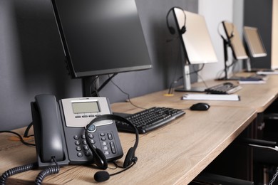 Photo of Stationary phone and headset near modern computer on wooden desk in office. Hotline service