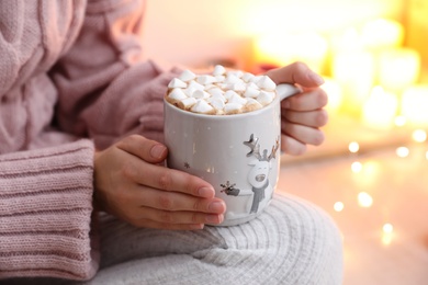 Photo of Woman holding cup of hot drink with marshmallows indoors, closeup. Magic Christmas atmosphere