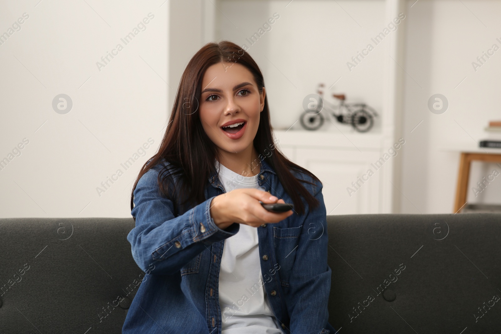 Photo of Beautiful woman changing TV channels with remote control on sofa at home