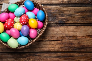 Photo of Colorful Easter eggs in wicker basket on wooden table, top view. Space for text