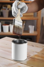Photo of Man dipping brush into can with varnish at wooden table indoors, closeup