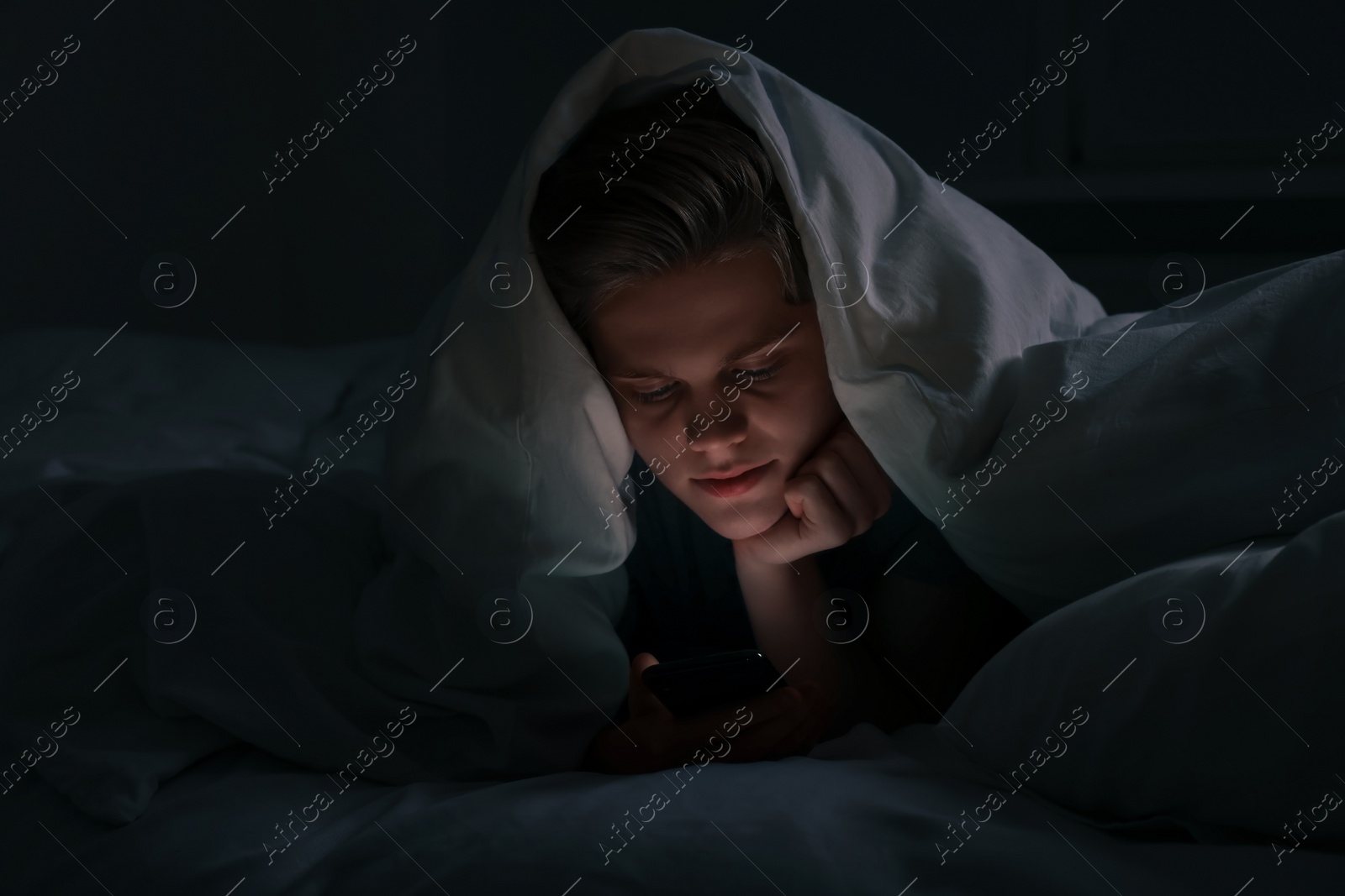 Photo of Teenage boy using smartphone under blanket on bed at night. Internet addiction