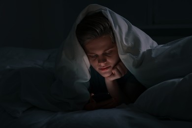 Photo of Teenage boy using smartphone under blanket on bed at night. Internet addiction