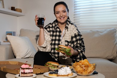 Happy overweight woman with glass of cola and burger on sofa at home
