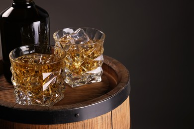 Whiskey with ice cubes in glasses and bottle on wooden barrel against dark background, space for text