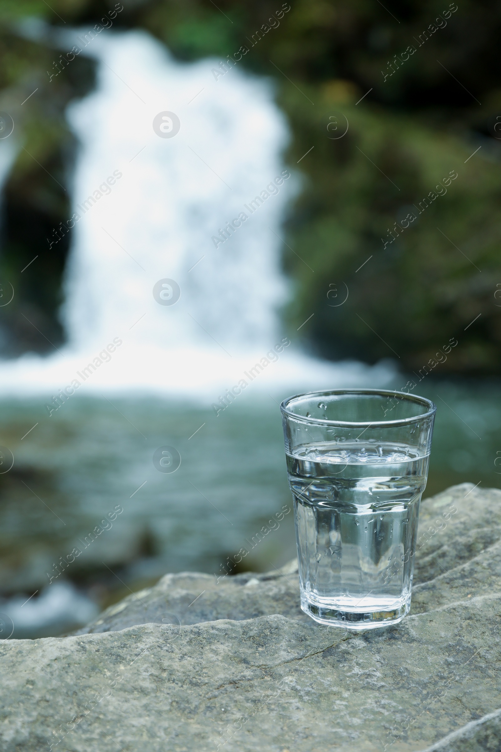 Photo of Glass of fresh water on stone near waterfall outdoors. Space for text