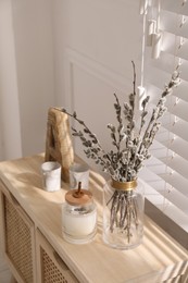 Photo of Glass vase with pussy willow tree branches and candle on wooden commode near window in room