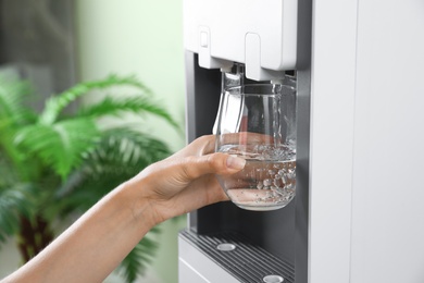 Photo of Woman filling glass from water cooler indoors, closeup. Refreshing drink